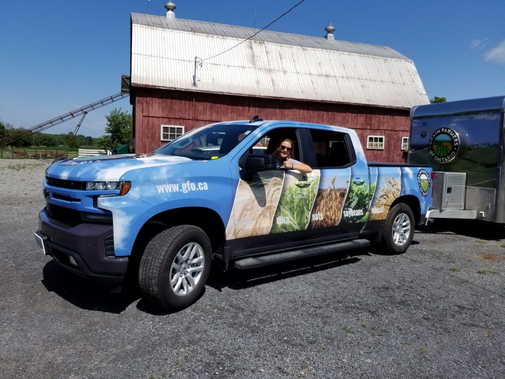 Photo of Grain Discovery Ambassador Driving Truck and Trailer with red barn in the background and Field Crop branded truck! Giving Thumbs Up