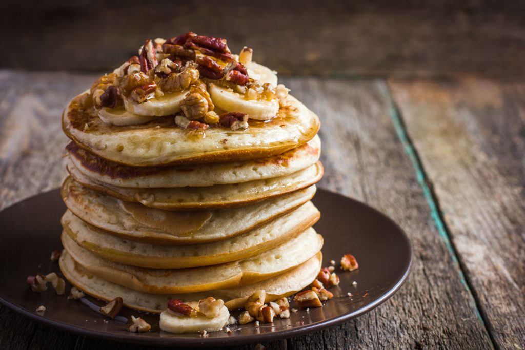 Plate of homemade whole wheat pancakes topped with bananas, walnuts, pecans and drizzled with maple syrup. 
