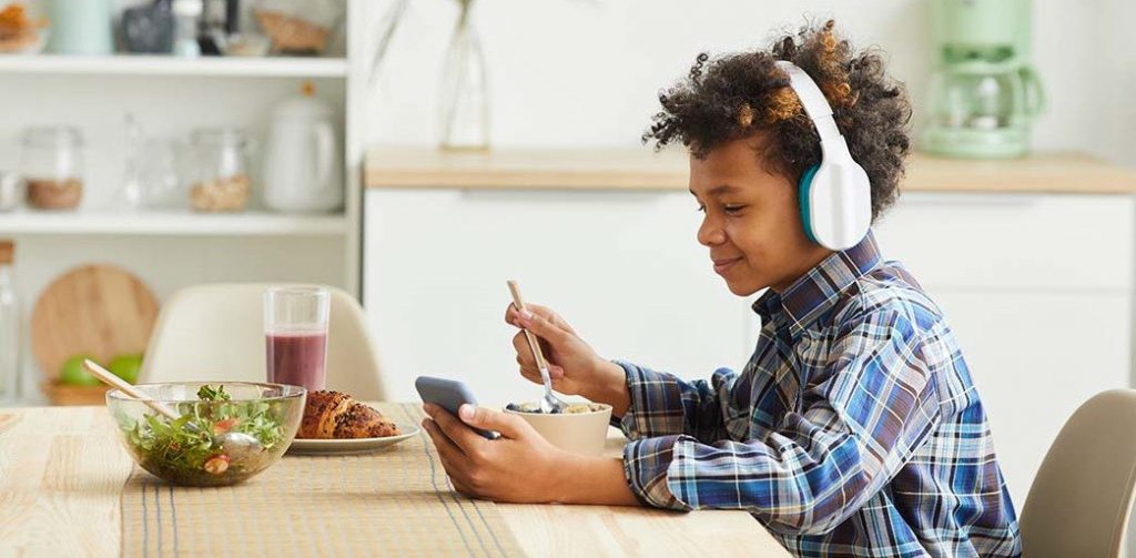 African boy in headphones using his mobile phone while eating breakfast in the kitchen