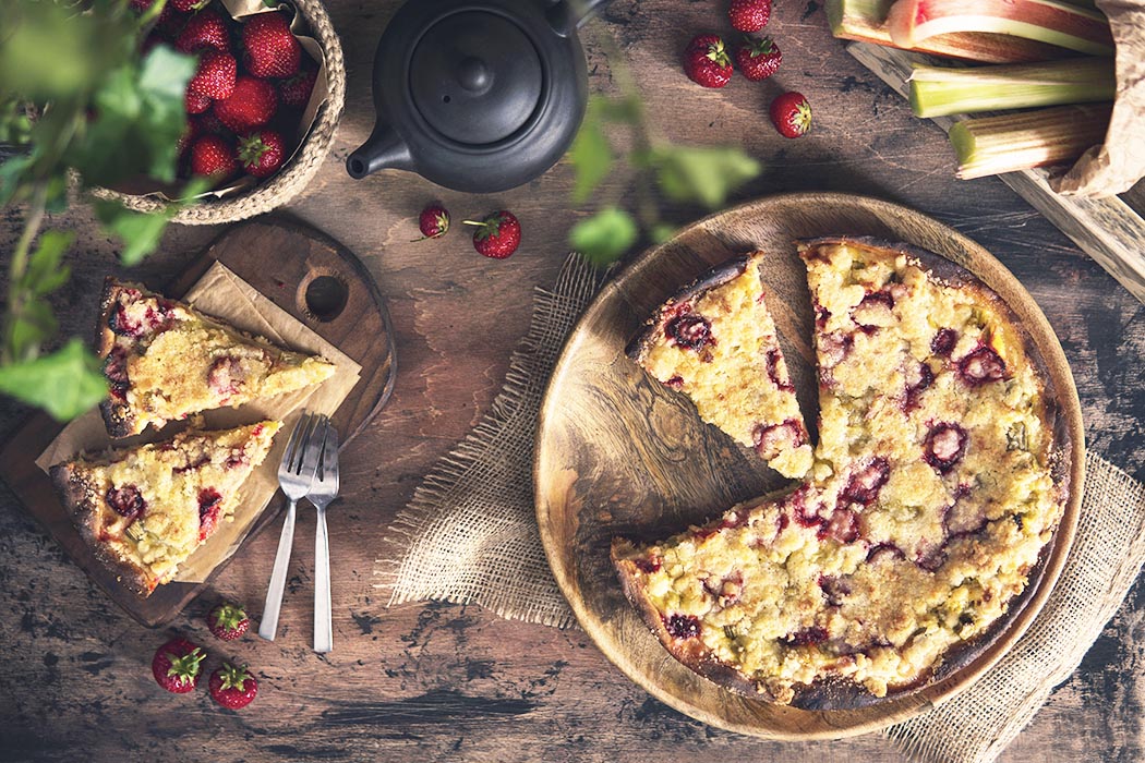 Rhubarb crumble pie with strawberry on rustic wooden table with fresh rhubarb, berries, plant. Beautiful still life homemade dessert. Top view, flat lay. Natural light at kitchen, organic products.